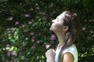 Woman closing her eyes in sunlight near purple flowers, practicing mindfulness and calm to ease overwhelm.