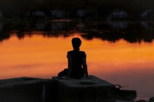 A silhouette of a person sitting peacefully by the lake which reflects stillness, calm, and peacefulness.
